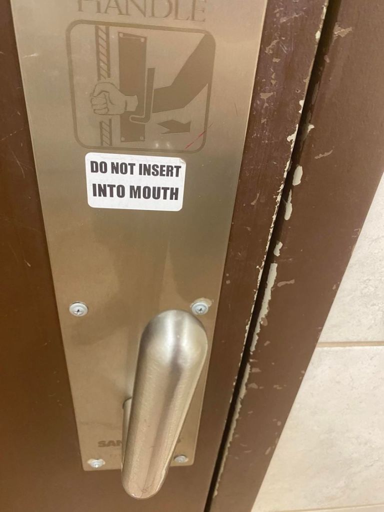 public restroom door handle. someone has added a sticker that says "do not insert into mouth."