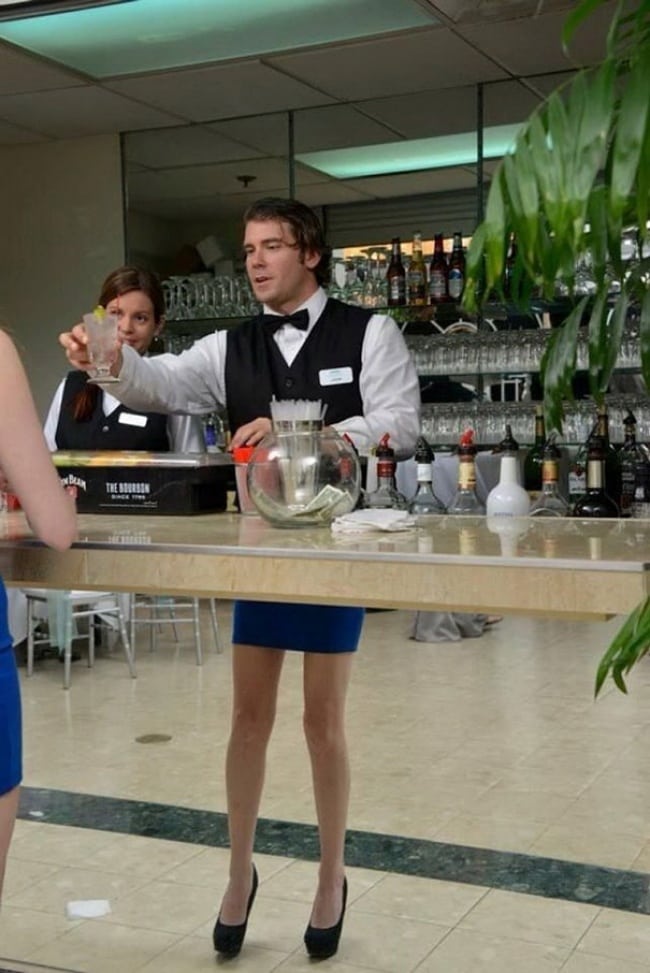 bartender standing behind reflective bar. Woman ordering drink wearing skirt and heels is reflected, so it looks like bartender is wearing skirt and heels.