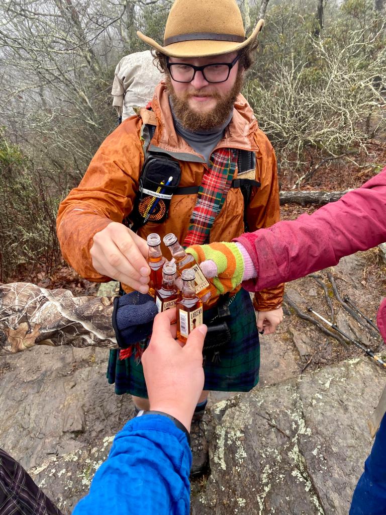 four hikers toasting with liquor bottles. One man is wearing camo so you can't see his arm in the picture
