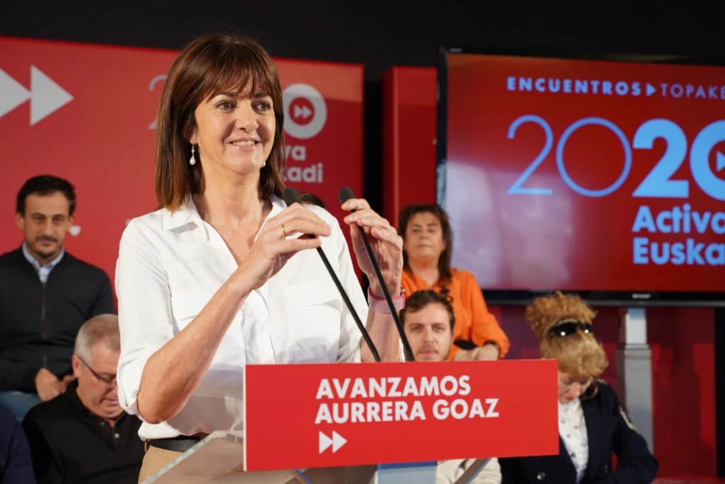 still from a video of politician giving a speech. Behind her, a woman looking down resembles Alf wearing sunglasses.
